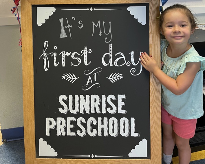 Child next to a blackboard that reads "It's My first day at SUNRISE PRESCHOOL"