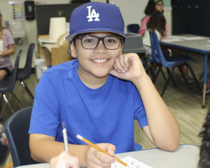 Child in glasses and blue LA hat smiling while writing at a desk