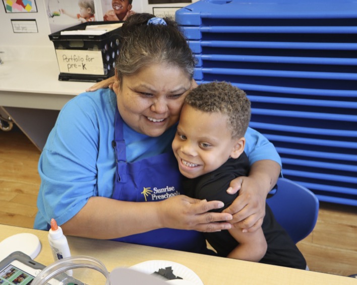 Person in blue apron hugging a child in a classroom