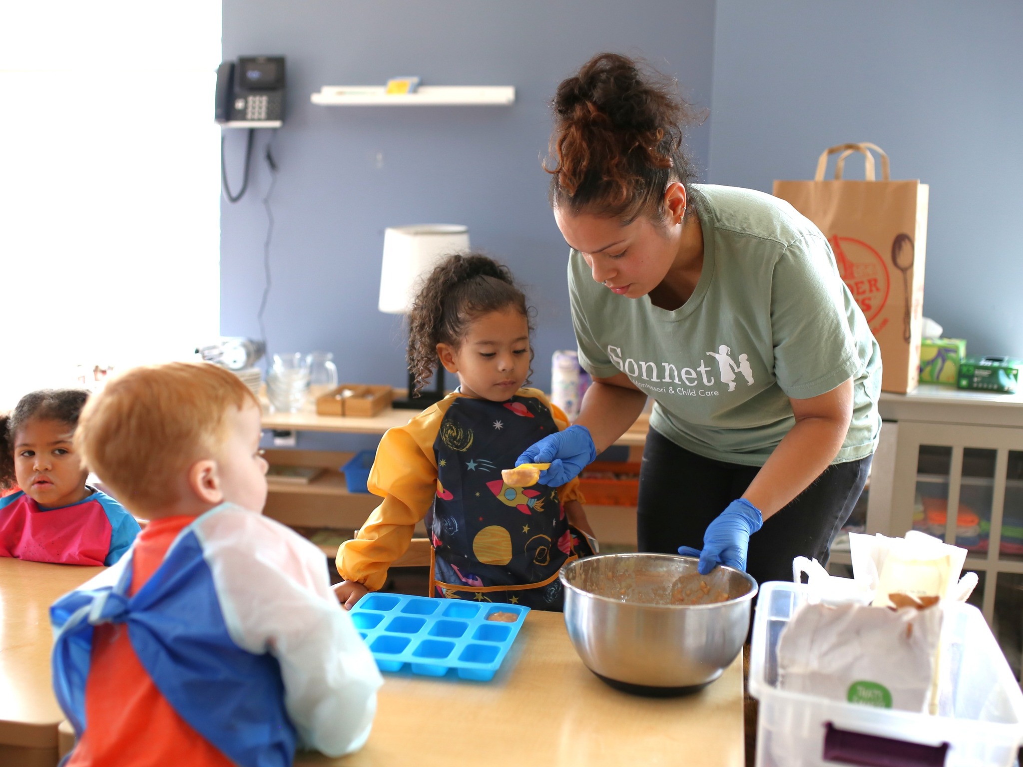 Adult helping child in smock put food into an ice cube tray in a classroom