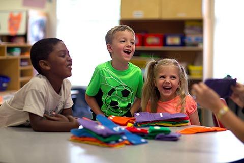 Three children laughing at a table with colorful craft supplies