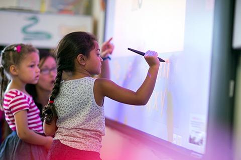 Child writing on an interactive whiteboard with a stylus, another child watching