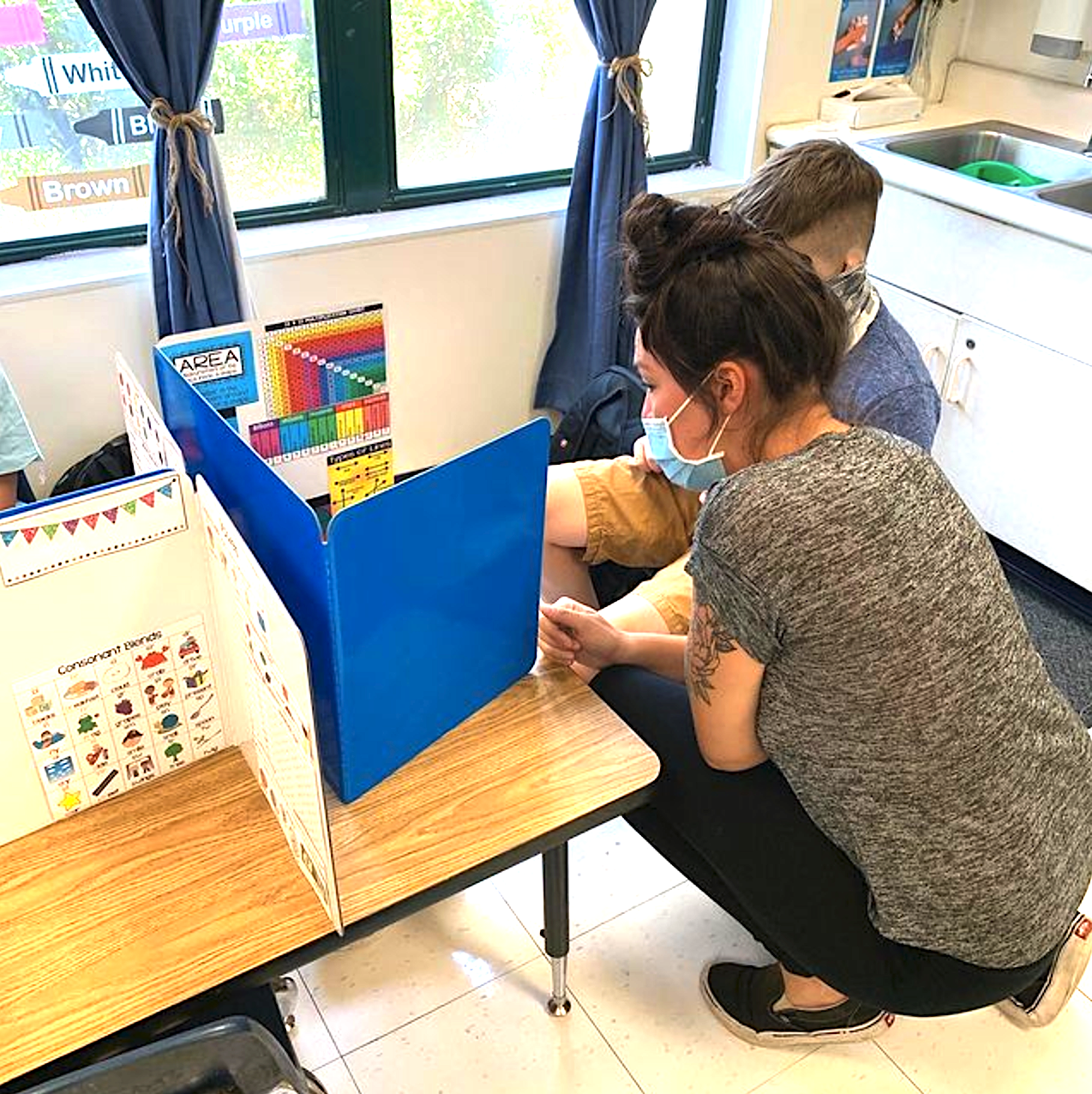 Person in mask crouching next to a child at a desk with a privacy divider