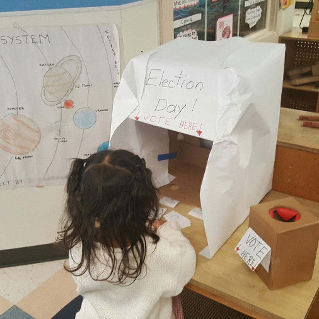 Child at a voting booth with "Election Day" and "Vote Here" signs