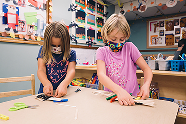 Two children wearing masks doing crafts at a table in a classroom