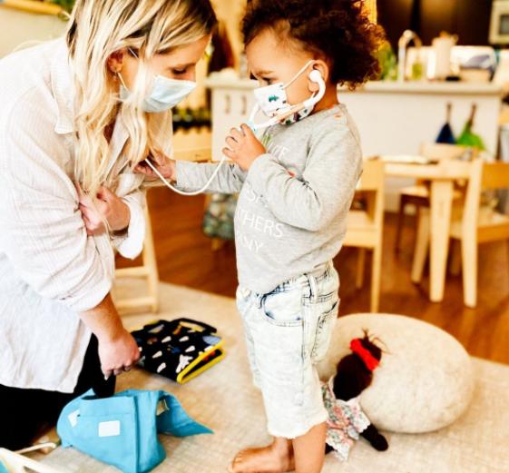 Person and child wearing masks, child holding stethoscope to person's chest